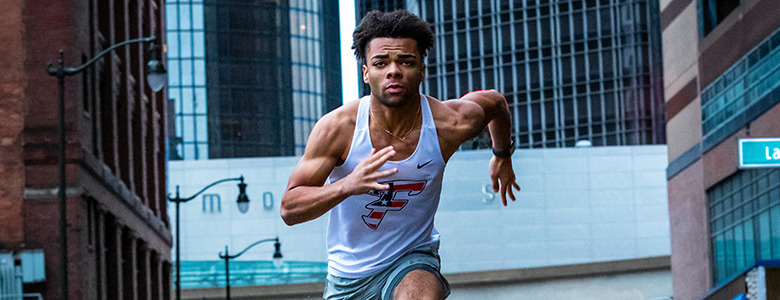 A high school senior in athletic gear sprints down a downtown Detroit street, perfect for vibrant senior pictures. He wears a white tank top with a logo and gray shorts. Tall buildings and a street sign labeled "Larned" are visible in the background.