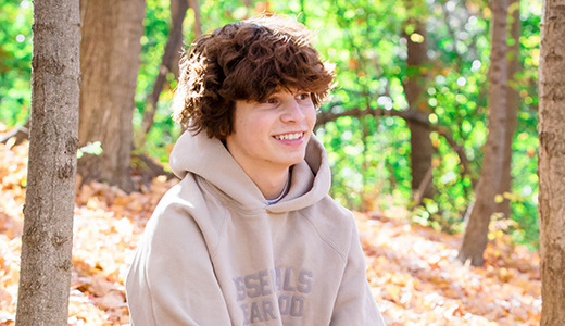 A high school senior with curly hair smiles during their fall senior portraits, seated in a sunlit, wooded area. They're wearing a beige hoodie as the ground is covered in autumn leaves, and the trees display vibrant green and yellow foliage.