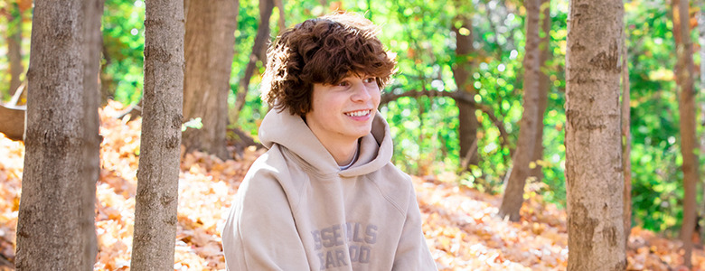 A high school senior with curly hair smiles during their fall senior portraits, seated in a sunlit, wooded area. They're wearing a beige hoodie as the ground is covered in autumn leaves, and the trees display vibrant green and yellow foliage.