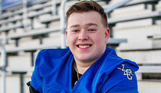 A high school senior wearing a blue football jersey sits on bleachers, smiling at the camera. The stands are empty, and the background is slightly blurred.
