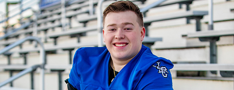 A high school senior wearing a blue football jersey sits on bleachers, smiling at the camera. The stands are empty, and the background is slightly blurred.