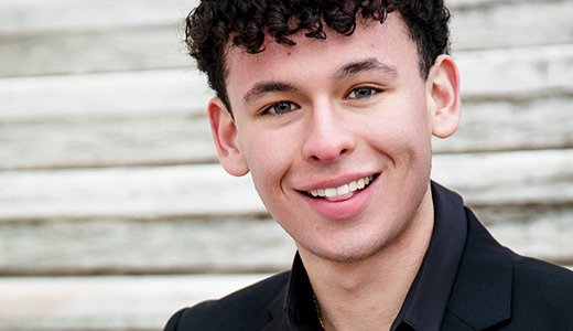 A high school senior with curly hair smiles while sitting on outdoor stone steps, capturing the essence of Macomb Senior Pictures. He is wearing a black blazer, perfectly blending sophistication with the natural charm of his surroundings.