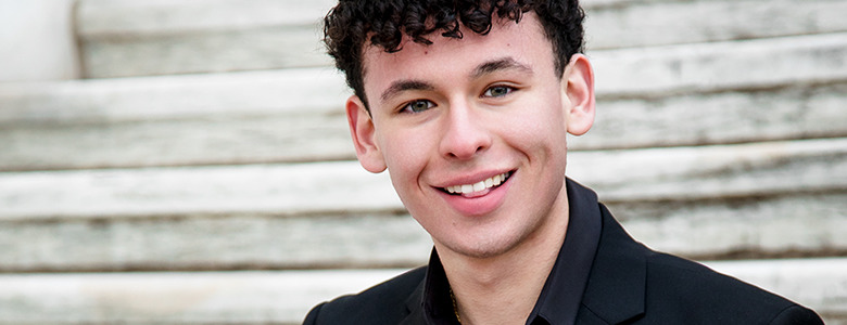 A high school senior with curly hair smiles while sitting on outdoor stone steps, capturing the essence of Macomb Senior Pictures. He is wearing a black blazer, perfectly blending sophistication with the natural charm of his surroundings.