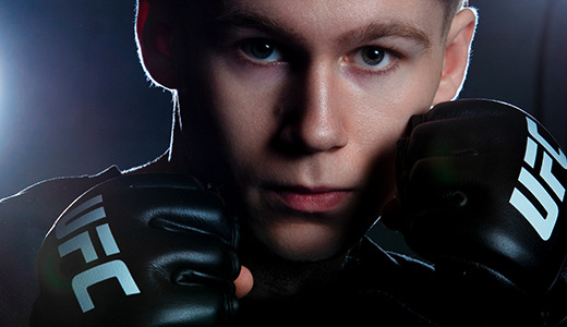 A high school senior with a serious expression, capturing the intensity of Ann Arbor senior portraits, wears UFC gloves and stands in a fighting stance. The background is dark with a bright light shining, creating a dramatic contrast.