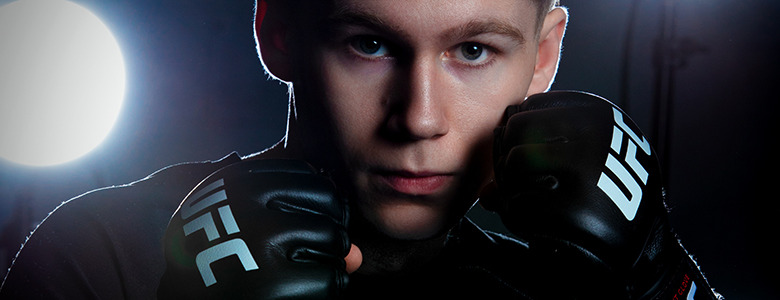 A high school senior with a serious expression, capturing the intensity of Ann Arbor senior portraits, wears UFC gloves and stands in a fighting stance. The background is dark with a bright light shining, creating a dramatic contrast.