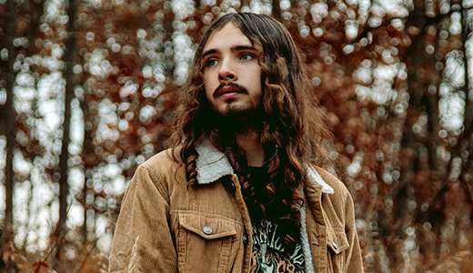 A high school senior with long hair and a beard stands in a forest, wearing a brown jacket over a patterned shirt. The background is filled with trees and autumn leaves, creating a warm, earthy atmosphere.