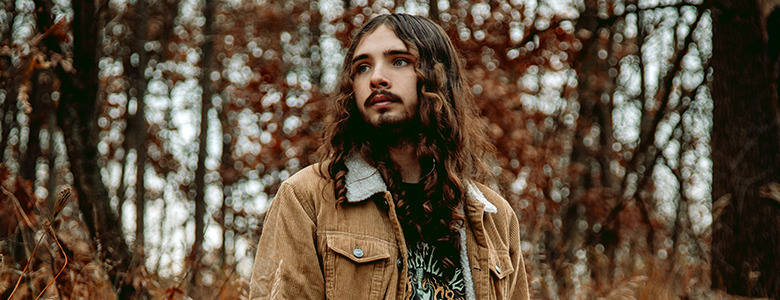 A high school senior with long hair and a beard stands in a forest, wearing a brown jacket over a patterned shirt. The background is filled with trees and autumn leaves, creating a warm, earthy atmosphere.