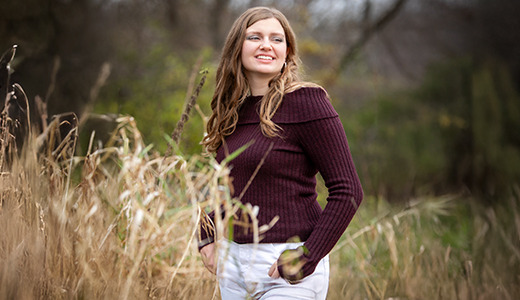 A high school senior stands in a field of tall grass, smiling and looking to her right during her Ypsilanti senior pictures. She is wearing a maroon sweater and light-colored pants. The background shows blurred trees and greenery, capturing the essence of this special moment.