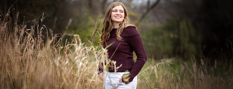 A high school senior stands in a field of tall grass, smiling and looking to her right during her Ypsilanti senior pictures. She is wearing a maroon sweater and light-colored pants. The background shows blurred trees and greenery, capturing the essence of this special moment.