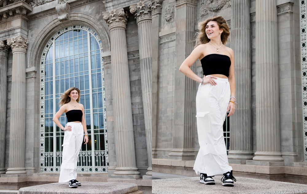 A high school senior with long hair stands in front of Michigan Central Station in Detroit, MI for her Detroit senior pictures session with Detroit photographer, Paul Manoian. She is wearing a black strapless top, white pants, and sneakers. She smiles confidently, posing with one hand on her hip.