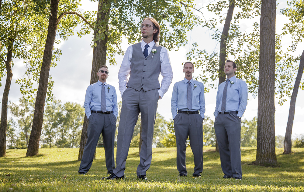 A groom in a gray suit stands confidently in a sunlit park, surrounded by three groomsmen in matching gray pants and light blue shirts. They are positioned among tall trees with a grassy field and clear sky in the background.