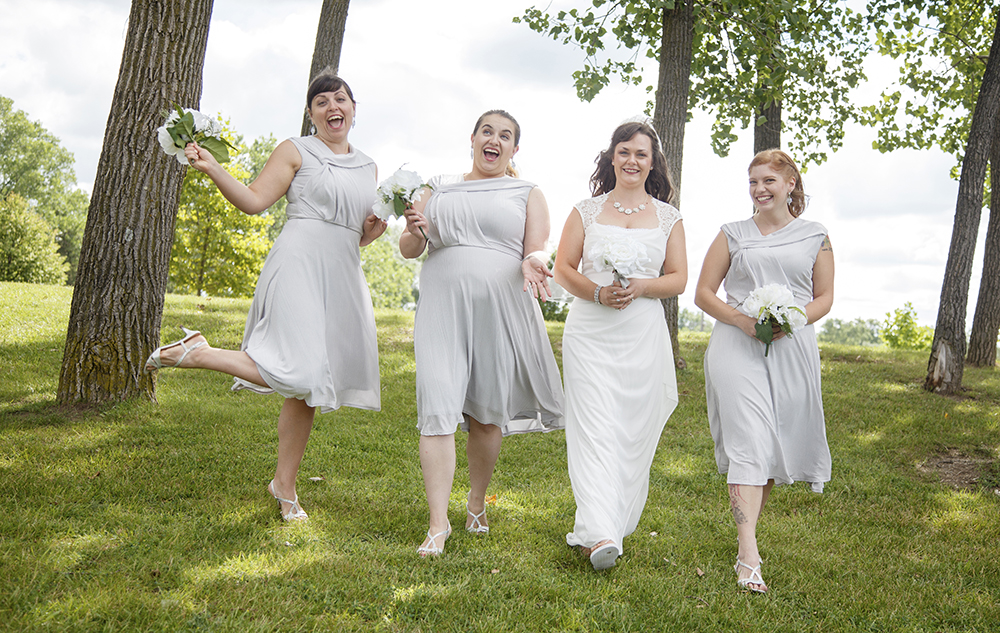 Four women are joyfully walking on grass under tall trees. Three bridesmaids in matching light gray dresses surround a bride in a white gown. All hold white bouquets, smiling brightly in a sunny outdoor setting.