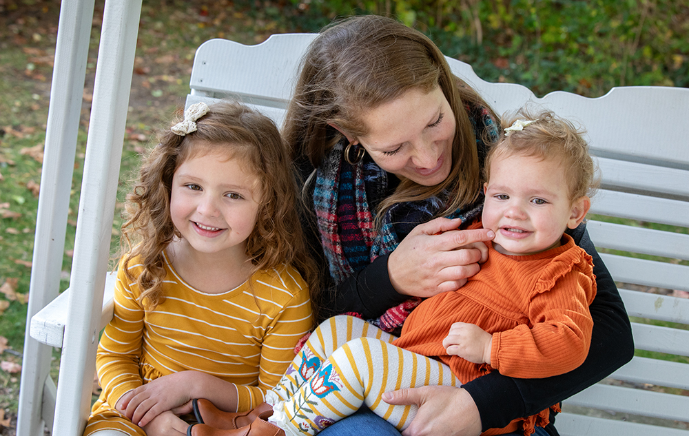 A woman sits on a porch swing with two young girls for a Livonia family portraits session with Livonia family photographer, Paul Manoian. The girls, wearing orange outfits, are smiling at the camera. The woman, wrapped in a colorful scarf, holds the younger child and smiles affectionately. Greenery is visible in the background.