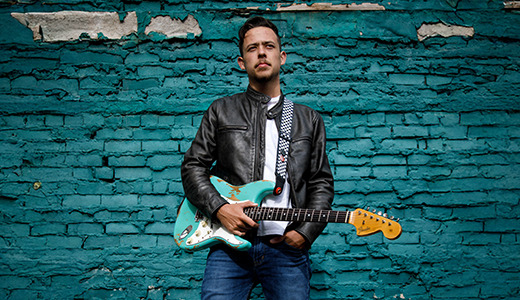 A high school senior wearing a leather jacket and jeans stands against a teal brick wall, holding an electric guitar for Rock n Roll senior pictures in Detroit, MI with Detroit photographer, Paul Manoian.
