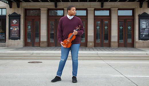 A high school senior stands on a street holding a violin, looking to the side. They are in front of a large building with wooden doors and decorative glass panels. The scene suggests a calm urban setting.