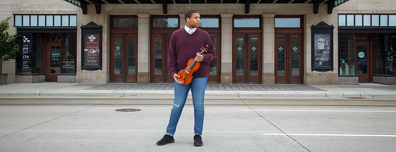 A high school senior stands on a street holding a violin, looking to the side. They are in front of a large building with wooden doors and decorative glass panels. The scene suggests a calm urban setting.