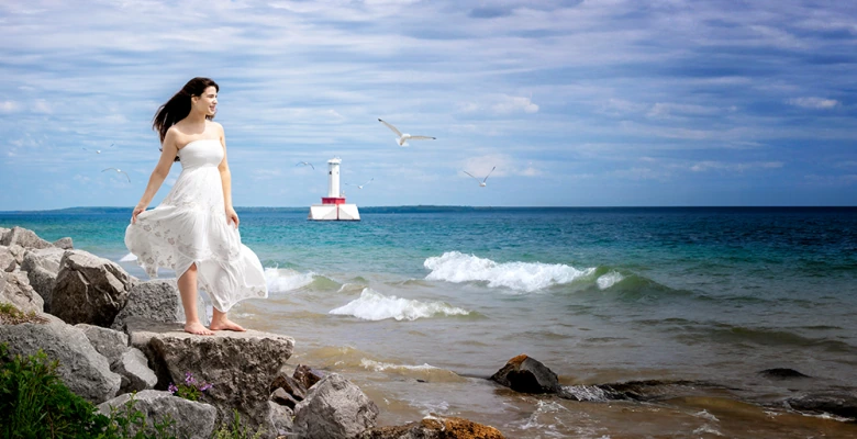 A high school senior girl in a flowing white dress stands on rocky terrain by a beach. She gazes towards the sea, with a lighthouse in the background and seagulls flying in the cloudy sky. Waves crash gently on the shore.