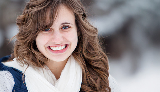 A high school senior girl with long wavy hair smiles warmly while standing outdoors, as if posing for senior pictures in the snow. Wrapped in a white scarf and blue jacket, they blend seamlessly into the winter scene of snow-covered trees.
