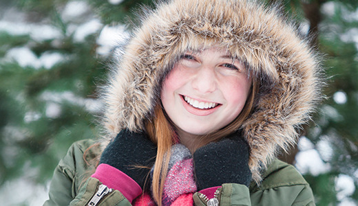 A high school senior smiling, wearing a fur-lined hooded green coat and dark gloves, stands outside on a snowy day. Snow-covered trees are in the background, creating a wintery scene perfect for capturing enchanting snow senior pictures.