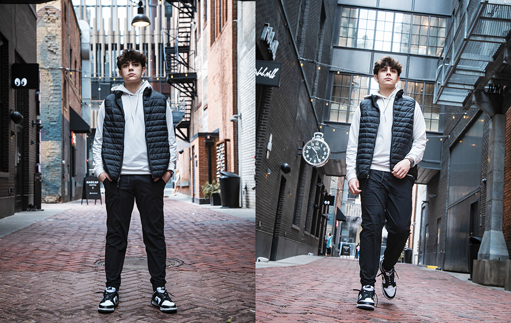 Senior Pictures Outfits Ideas for Guys in 2025 Senior Pictures Outfits Ideas for Guys in 2026: A high school senior boy in a black puffer vest, white hoodie, and black pants is standing and walking in an urban alley. The setting features brick and modern architecture, with outdoor lights and an overhead clock on the brick wall.