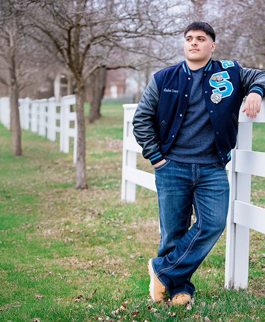 A young man in a letterman jacket leans on a white wooden fence in a grassy, tree-lined outdoor setting, with bare trees and a pathway visible in the background.