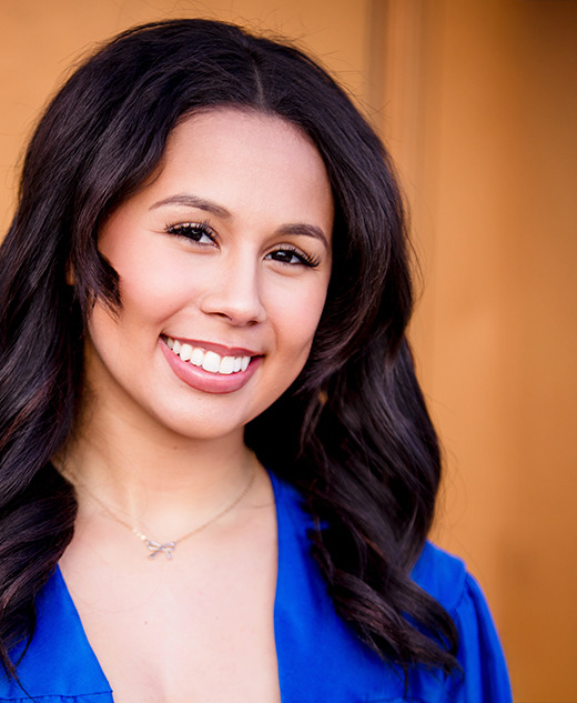 A high school senior girl with long dark hair smiles, wearing a blue graduation gown and a necklace, standing in front of a blurred golden-brown background—an example of elegant senior portraits.