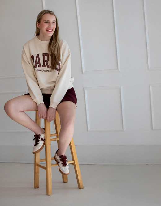 A smiling girl sits on a wooden stool in a minimal white room, wearing a cream "PARIS" sweatshirt, maroon shorts, and matching sneakers—capturing the relaxed vibe often seen in kids headshots.