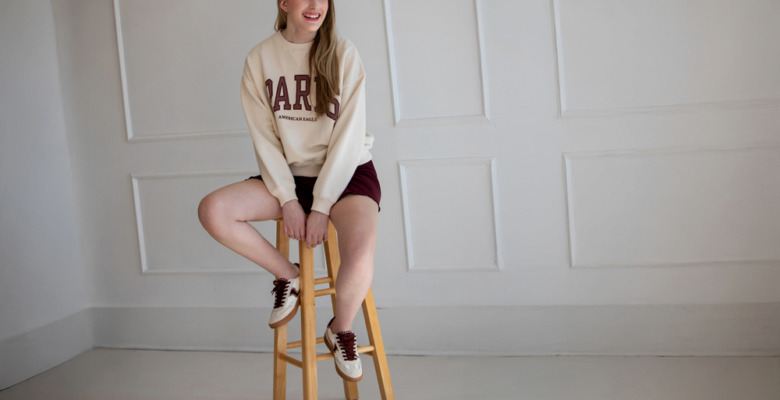 A smiling girl sits on a wooden stool in a minimal white room, wearing a cream "PARIS" sweatshirt, maroon shorts, and matching sneakers—capturing the relaxed vibe often seen in kids headshots.