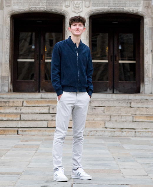 A high school senior boy stands on a stone pathway in front of a large, ornate building with double wooden doors and decorative lanterns—hands in pockets, smiling at the camera—perfect for casual senior portraits in Ann Arbor.
