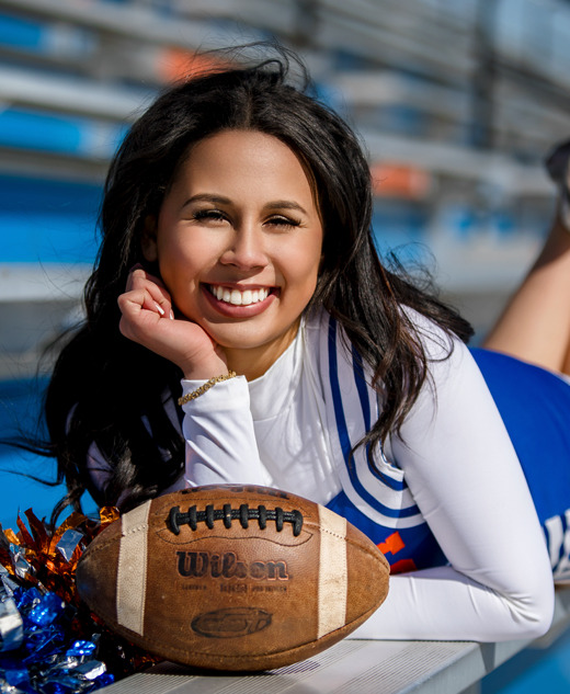 A high school senior cheerleader in a blue and white uniform smiles while lying on bleachers, resting her head on her hand. Capturing the spirit of senior pictures, a football and pom-poms sit before her. The bleachers and sky form a picturesque backdrop.