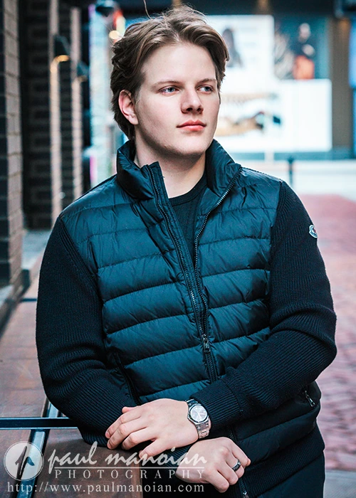A high school senior guy wearing a black quilted vest and sweater leans casually against a railing, embodying Detroit urban chic. He's looking off to the side with a thoughtful expression. Photographed by Paul Manoian Photography.