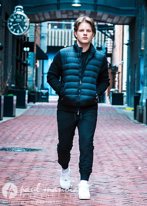 A high school senior walks confidently down a cobblestone street lined with brick buildings, embodying Detroit urban chic in a black puffer jacket, dark pants, and white sneakers. Their hands are in their pockets as a large clock ticks in the background.