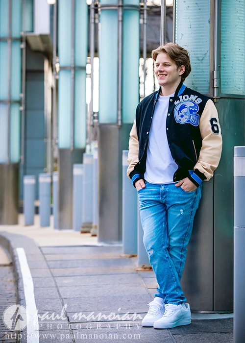 A high school senior guy with light brown hair, embodying Detroit urban chic in a black and beige varsity jacket, blue jeans, and white sneakers, stands casually against a pillar on a pathway lined with teal glass panels.