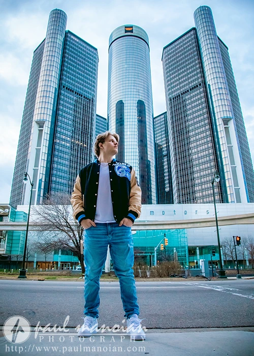 A high school senior guy exudes Detroit urban chic as they confidently stand before the Renaissance Center skyscrapers. Clad in a brown and black jacket and jeans, they gaze slightly upwards. The cloudy sky looms overhead while bare trees accentuate the city's backdrop.