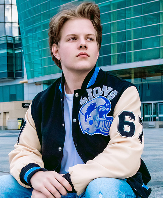 High school senior guy wearing a Detroit Lions varsity jacket with the number 65, sitting in front of the Renaissance Center building in downtown Detroit, MI for his senior pictures session with award-winning senior photographer, Paul Manoian. He has light brown hair and are looking to the side, with a thoughtful expression.