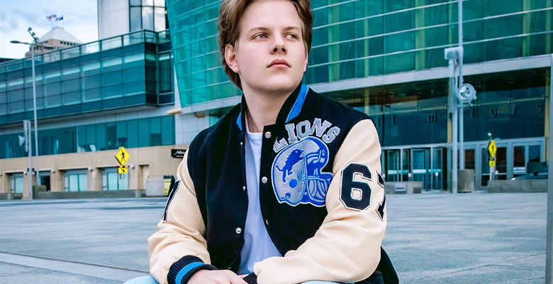 High school senior guy wearing a Detroit Lions varsity jacket with the number 65, sitting in front of the Renaissance Center building in downtown Detroit, MI for his senior pictures session with award-winning senior photographer, Paul Manoian. He has light brown hair and are looking to the side, with a thoughtful expression.
