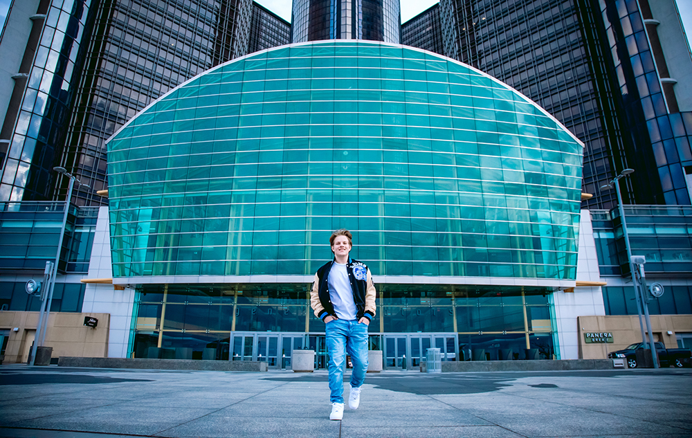 A person in a jacket and jeans stands confidently in front of a large, modern building with a blue-green glass facade and multiple stories. The building's architecture features geometric lines and reflective surfaces.