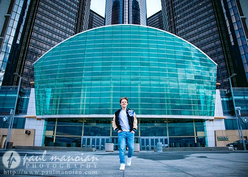 A high school senior guy wearing a casual jacket and jeans exudes Detroit urban chic as they walk confidently in front of a modern glass building with a curved facade. The building towers above, featuring reflective windows and metal accents.