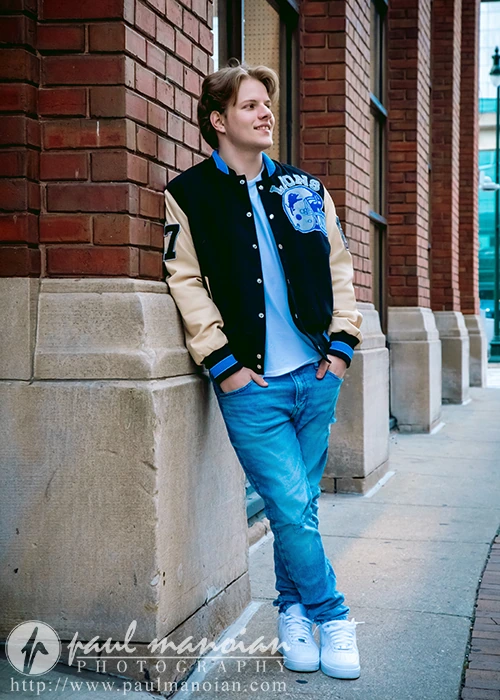 A high school senior guy embodying Detroit urban chic in a varsity jacket, jeans, and white sneakers leans against a brick wall on a city sidewalk, smiling and gazing into the distance.
