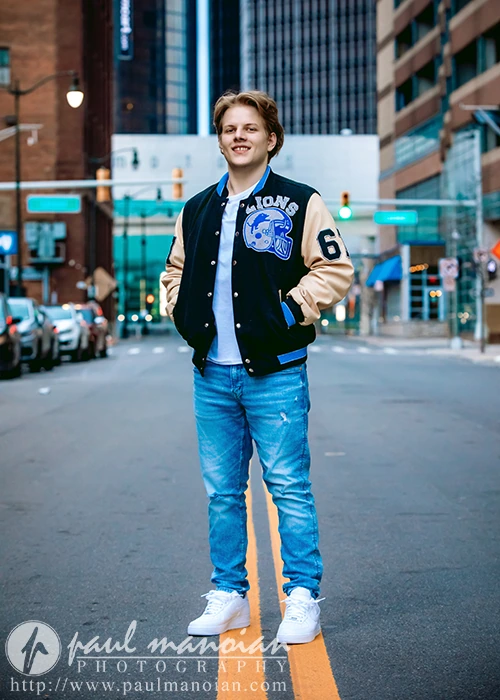 A high school senior guy stands confidently on a Detroit street, embodying Detroit urban chic. They wear a varsity jacket with "Lions" lettering, jeans, and white sneakers. Tall buildings and traffic lights frame the vibrant city backdrop.