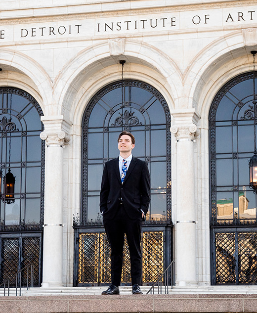 A person in a suit stands on the steps in front of the Detroit Institute of Arts, with three large arched windows and the building’s name visible above, capturing stylish modern senior portraits.