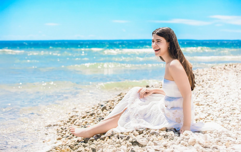 A high school senior girl in a white dress sits on a rocky beach at Mackinac Island, smiling joyfully under a bright blue sky. The clear ocean waves gently lap against the shore, creating a serene and cheerful atmosphere perfect for senior pictures.