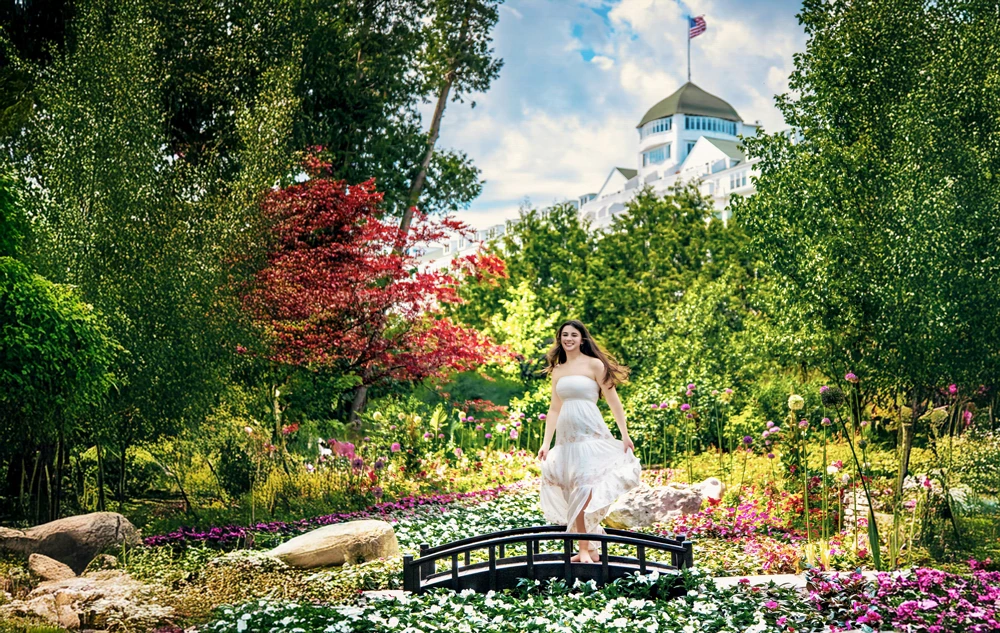 A high school senior girl in a white dress poses gracefully on a small, arching bridge in a vibrant garden on Mackinac Island, capturing timeless senior pictures. Behind her, a large white building with a flag atop rises against a partly cloudy sky, complementing the lush greenery and colorful blossoms.