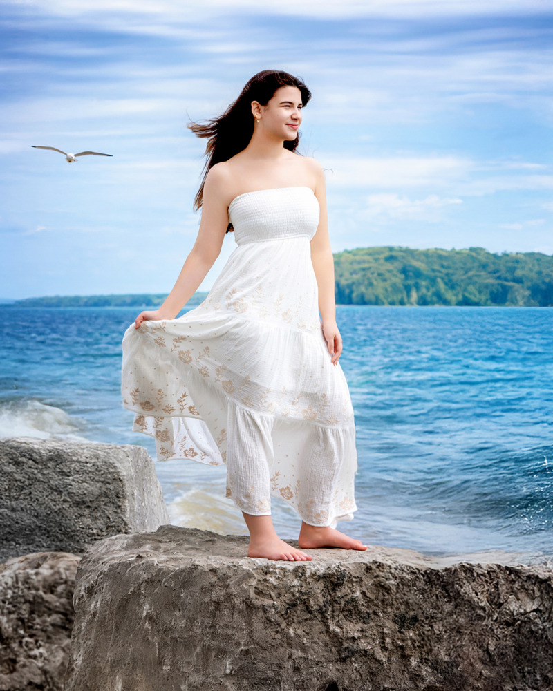 A high school senior girl in a white dress stands barefoot on Mackinac Island's rocky shore, holding her dress gently. The ocean and sky create a serene backdrop, with a seagull flying nearby. Lush green hills are visible in the distance, making it an ideal spot for senior pictures.