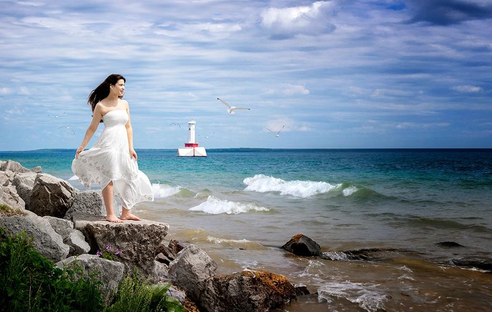 A high school senior girl in a white dress stands on rocky terrain by the ocean, gazing out at the water, reminiscent of Mackinac Island senior pictures. Seagulls fly around, and a lighthouse is visible in the distance beneath a cloudy blue sky. Waves crash gently against the rocks.