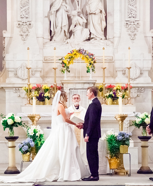 A bride and groom stand holding hands at the altar in a church, facing each other. An officiant stands between them. Two attendants stand on either side, as a Detroit wedding photographer captures the flowers and candlelit decor.