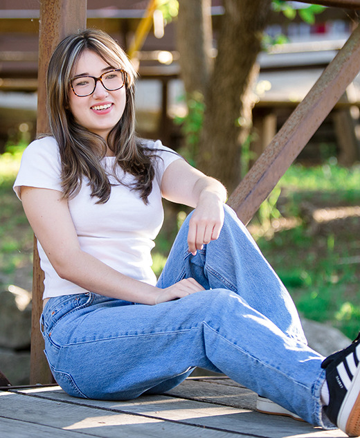 A high school senior girl with long brown hair, glasses, a white t-shirt, and blue jeans sits smiling on a wooden bridge outdoors, surrounded by greenery and sunlight at Franklin Cider Mill.