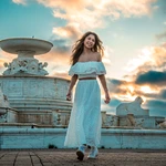 A high school senior girl in a white off-shoulder top and long skirt walks and smiles in front of a large ornate fountain at sunset, with dramatic clouds and warm sunlight in the sky behind her.