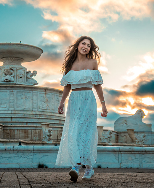 A high school senior girl in a white off-shoulder top and long skirt walks and smiles in front of a large ornate fountain at sunset, with dramatic clouds and warm sunlight in the sky behind her.
