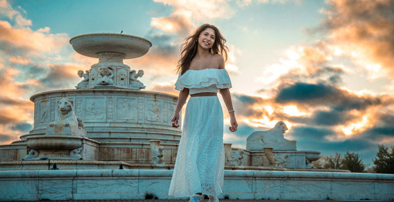 A high school senior girl in a white off-shoulder top and long skirt walks and smiles in front of a large ornate fountain at sunset, with dramatic clouds and warm sunlight in the sky behind her.